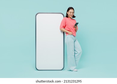 Joven cuerpo entero sonriendo feliz mujer de etnia asiática de 20 años en un puesto de suéter rosa cerca de un gran teléfono celular con área de trabajo de pantalla en blanco charlando aislado en un fondo azul claro pastel.