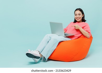 Full Body Young Smiling Happy Woman Of Asian Ethnicity 20s Wear Pink Sweater Sit In Bag Chair Hold Use Work Point Finger Oon Laptop Pc Computer Isolated On Pastel Plain Light Blue Background Studio.