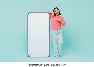 Full body young smiling happy woman of Asian ethnicity 20s in pink sweater point finger on big mobile cell phone with blank screen workspace area isolated on pastel plain light blue background studio - Shutterstock ID 2133623459