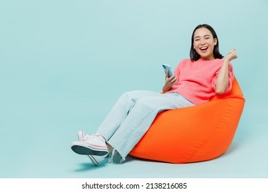 Full body young smiling happy woman of Asian ethnicity 20s wearing pink sweater sit in bag chair hold in hand use mobile cell phone do winner gesture isolated on pastel plain light blue background.