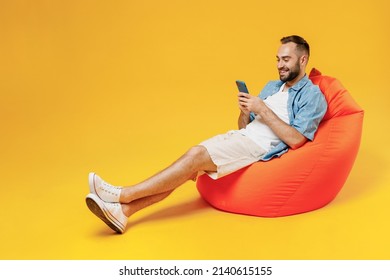 Full body young smiling happy satisfied man 20s wearing blue shirt white t-shirt sit in bag chair use mobile cell phone isolated on plain yellow background studio portrait. People lifestyle concept. - Shutterstock ID 2140615155