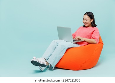 Full Body Young Smiling Happy Woman Of Asian Ethnicity 20s Wear Pink Sweater Sit In Bag Chair Hold Use Work On Laptop Pc Computer Isolated On Pastel Plain Light Blue Color Background Studio Portrait.