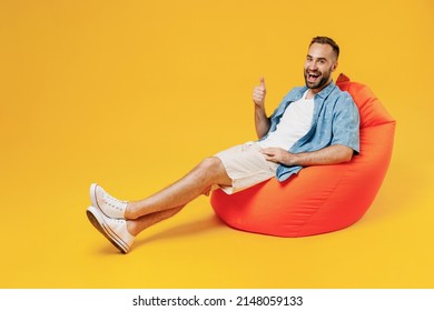 Full body young smiling happy cheerful man 20s wearing blue shirt white t-shirt sit in bag chair show thumb up gesture isolated on plain yellow background studio portrait. People lifestyle concept.