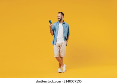 Full Body Young Smiling Happy Cool Caucasian Man 20s Wearing Blue Shirt White T-shirt Hold In Hand Use Mobile Cell Phone Isolated On Plain Yellow Background Studio Portrait. People Lifestyle Concept