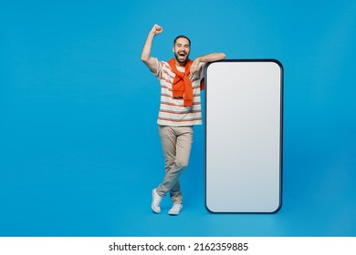 Full Body Young Smiling Happy Man 20s In Orange Striped T-shirt Stand Near Big Blank Screen Mobile Cell Phone With Workspace Copy Space Mockup Area Do Winner Gesture Isolated On Plain Blue Background