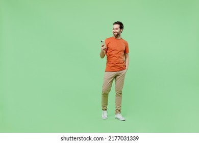 Full body young smiling happy fun man 20s wear casual orange t-shirt hold in hand use mobile cell phone isolated on plain pastel light green color background studio portrait. People lifestyle concept