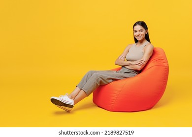 Full Body Young Smiling Happy Fun Cool Latin Woman 30s She Wear Basic Beige Tank Shirt Sit In Bag Chair Look Camera Isolated On Plain Yellow Color Backround Studio Portrait. People Lifestyle Concept