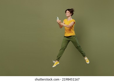 Full Body Young Smiling Happy Woman She 20s Wear Yellow T-shirt Jump High Run Hold In Hand Use Mobile Cell Phone Isolated On Plain Olive Green Khaki Background Studio Portrait People Lifestyle Concept