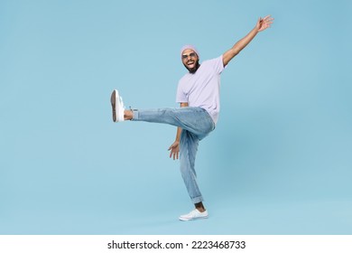 Full body young smiling happy man of African American ethnicity 20s wearing violet t-shirt hat glasses standing with raised up leg outstretched hands isolated on pastel blue background studio portrait