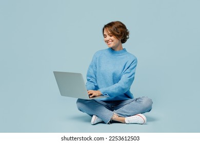 Full body young smiling happy fun cool IT woman wear knitted sweater hold use work on laptop pc computer isolated on plain pastel light blue cyan background studio portrait. People lifestyle concept