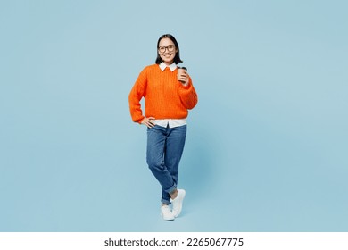 Full body young smiling happy woman of Asian ethnicity wear orange sweater glasses hold takeaway delivery craft paper brown cup coffee to go isolated on plain pastel light blue cyan background studio