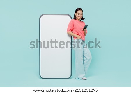 Photo of Full body young smiling happy woman of Asian ethnicity 20s in pink sweater stand near big mobile cell phone with blank screen workspace area chatting isolated on pastel plain light blue background.