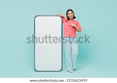Full body young smiling happy woman of Asian ethnicity 20s in pink sweater point finger on big mobile cell phone with blank screen workspace area isolated on pastel plain light blue background studio