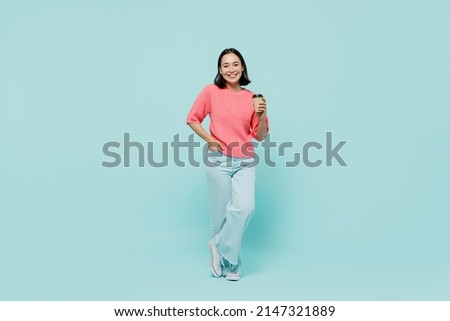 Full body young smiling happy woman of Asian ethnicity 20s wearing pink sweater hold takeaway delivery craft paper brown cup coffee to go isolated on pastel plain light blue color background studio