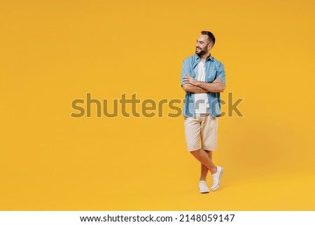 Full body young smiling happy fun minded cool man 20s wearing blue shirt white t-shirt hold hands crossed folded looking aside on copy space area isolated on plain yellow background studio portrait.