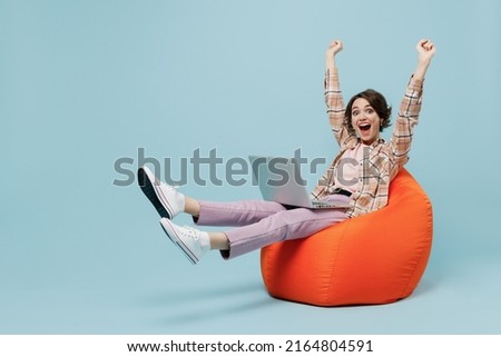 Full body young smiling happy woman 20s in brown shirt sit in bag chair with outstretched hands finish job hold use work on laptop pc computer isolated on pastel plain light blue background studio.
