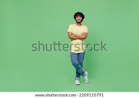 Full body young smiling happy Indian man 20s in basic yellow t-shirt hold hands crossed folded look camera isolated on plain pastel light green background studio portrait. People lifestyle concept.