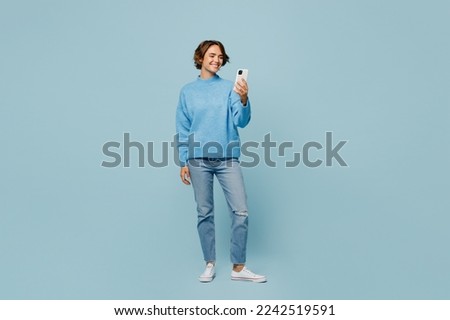 Full body young smiling happy woman wear knitted sweater look camera hold in hand use mobile cell phone isolated on plain pastel light blue cyan background studio portrait. People lifestyle concept