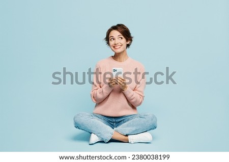 Full body young smiling happy cheerful woman she wear beige knitted sweater casual clothes sit hold in hand use mobile cell phone isolated on plain pastel light blue cyan background. Lifestyle concept