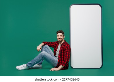 Full Body Young Smiling Man He 20s Wear Red Shirt Grey T-shirt Sit Big Huge Blank Screen Mobile Cell Phone With Workspace Copy Space Mockup Area Look Camera Isolated On Plain Dark Green Background.