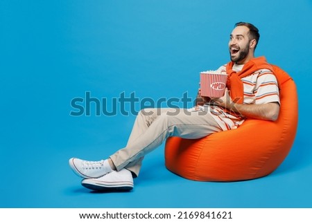 Full body young smiling man 20s in orange striped t-shirt sit in bag chair hold takeaway bucket of popcorn watch movie film isolated on plain blue background studio portrait. People lifestyle concept