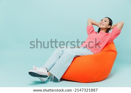 Full body young smiling minded fun woman of Asian ethnicity 20s wear pink sweater sit in bag chair hold hands behind neck isolated on pastel plain light blue color background People lifestyle concept