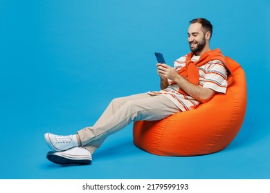 Full Body Young Smiling Satisfied Fun Happy Man 20s Wear Orange Striped T-shirt Sit In Bag Chair Hold In Hand Use Mobile Cell Phone Isolated On Plain Blue Background Studio. People Lifestyle Concept