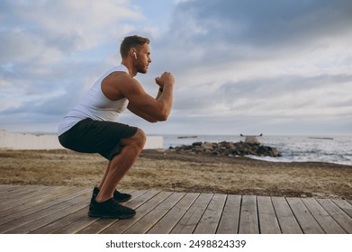 Full body young strong sporty athletic toned fit sportsman man wears sports clothes earphones do squats warm up train at sunrise sun dawn over sea beach outdoor on pier seaside in summer day morning - Powered by Shutterstock