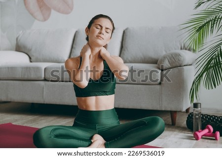 Full body young strong sporty athletic fitness trainer instructor woman wear green tracksuit sit on yoga mat suffering from pain spasm neck sprain train do exercises at home gym Workout sport concept