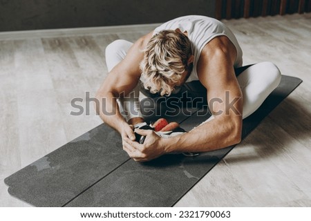 Full body young strong sporty sportsman man wear white tank shirt black shorts do stretch exercise for legs, sit at floor on yoga mat ean forward warm up training indoor at gym. Workout sport concept