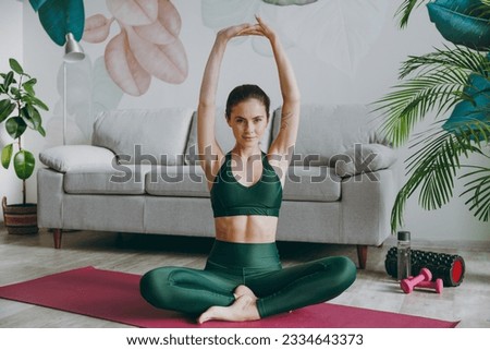 Full body young strong sporty athletic fitness trainer instructor woman wear green tracksuit sit on yoga mat raise up hands training do exercises at home gym indoor Workout sport motivation concept