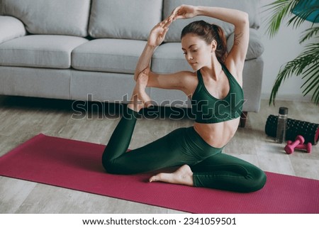 Full body young strong sporty athletic fitness trainer instructor woman wearing green tracksuit training doing stretching hand leg exercises for flexibility at home gym indoor. Workout sport concept