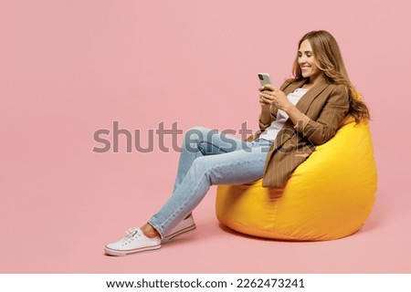 Full body young successful employee business woman 30s she wear casual brown classic jacket sit in bag chair hold in hand use mobile cell phone isolated on plain pastel light pink background studio