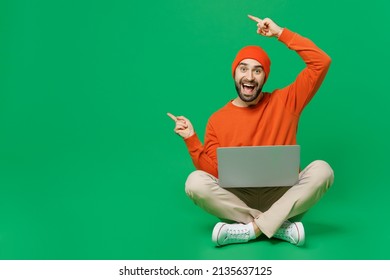 Full Body Young Surprised Amazed Happy Man 20s In Orange Sweatshirt Hat Sit Hold Use Work On Laptop Pc Computer Poin Index Finger Aside On Workspace Isolated On Plain Green Background Studio Portrait.