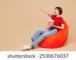 Full body young surprised happy woman wearing red t-shirt casual clothes sit in bag chair eat popcorn watch movie film point finger aside isolated on plain pastel beige background. Lifestyle concept