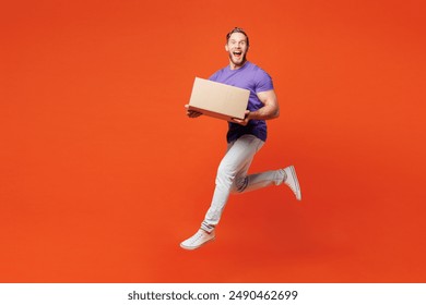 Full body young surprised shocked happy man he wearing purple t-shirt casual clothes holding cardboard box jump high hurry up isolated on red orange wall background studio portrait. Lifestyle concept