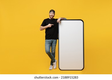 Full Body Young Tattooed Man He Wears Black T-shirt Cap Point Index Finger On Big Huge Blank Screen Mobile Cell Phone Workspace Mockup Area Isolated On Plain Yellow Background Tattoo Translate Fun.