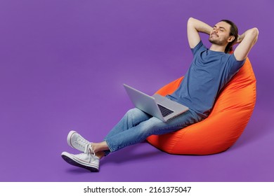 Full Body Young Tired Man 20s He Wear Basic Blue T-shirt Sit In Bag Chair Hold Use Work On Laptop Pc Computer Holding Hands Behind Neck Rest Isolated On Plain Purple Color Background Studio Portrait.