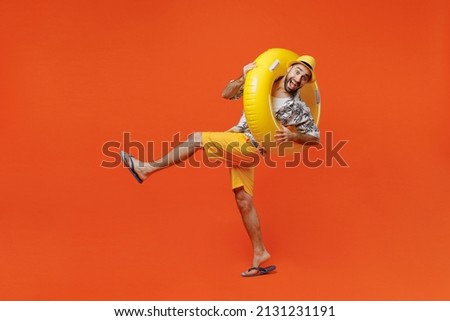 Full body young tourist man wear beach shirt hat hold inflatable ring lean back raise up leg fooling around isolated on plain orange background studio portrait Summer vacation sea rest sun tan concept