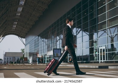 Full Body Young Traveler Serious Businessman Man 20s Wear Black Dinner Suit Walk Standing Outside At International Airport Terminal With Suitcase Valise Crossing Road Air Flight Business Trip Concept.