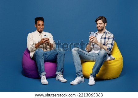 Full body young two friends happy men 20s wear white casual shirts together sit in bag chair hold in hand use mobile cell phone isolated plain dark royal navy blue background. People lifestyle concept