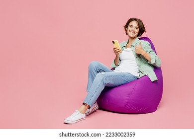 Full body young woman 20s she wear green shirt white t-shirt sit in bag chair hold in hand use mobile cell phone show thumb up isolated on plain pastel light pink background. People lifestyle concept - Powered by Shutterstock