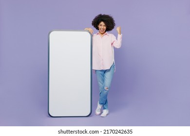 Full body young woman of African American ethnicity wear pink striped shirt stand near mobile cell phone with blank screen workspace area do winner gesture isolated on plain pastel purple background - Shutterstock ID 2170242635