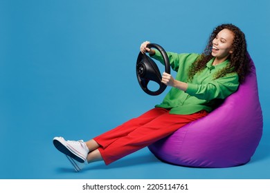 Full Body Young Woman Of African American Ethnicity 20s She Wear Green Shirt Sit In Bag Chair Hold Steering Wheel Pretending To Drive Car Isolated On Plain Blue Background. People Lifestyle Concept