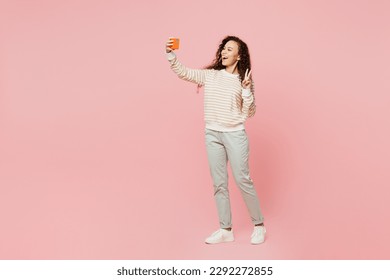 Full body young woman of African American ethnicity wear light casual clothes doing selfie shot on mobile cell phone post photo on social network show v-sign isolated on plain pastel pink background