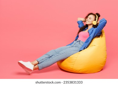 Full body young woman of African American ethnicity she wear blue shirt casual clothes sit in bag chair listen to music in headphones isolated on plain pastel pink background studio. Lifestyle concept - Powered by Shutterstock