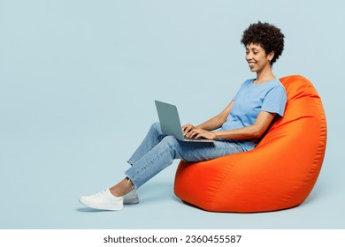 Full body young IT woman of African American ethnicity wear t-shirt casual clothes sit in bag chair hold use work on laptop pc computer isolated on plain pastel blue cyan background. Lifestyle concept - Powered by Shutterstock
