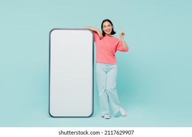 Full body young woman of Asian ethnicity in pink sweater stand near big mobile cell phone with blank screen workspace area point finger up with new idea isolated on pastel plain light blue background - Shutterstock ID 2117629907
