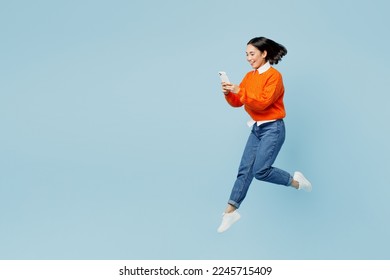 Full body young woman of Asian ethnicity wear orange sweater glasses jump high hold in hand use mobile cell phone isolated on plain pastel light blue cyan background studio. People lifestyle concept - Shutterstock ID 2245715409