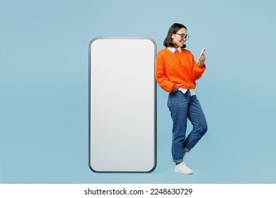 Full body young woman of Asian ethnicity wear orange sweater glasses big huge blank screen mobile cell phone with area use smartphone chat online isolated on plain pastel light blue cyan background - Powered by Shutterstock